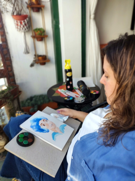 Adriana Fresnedo trabajando en un retrato en acuarela de cabello azul, sentada al aire libre con pinturas y marcadores cerca